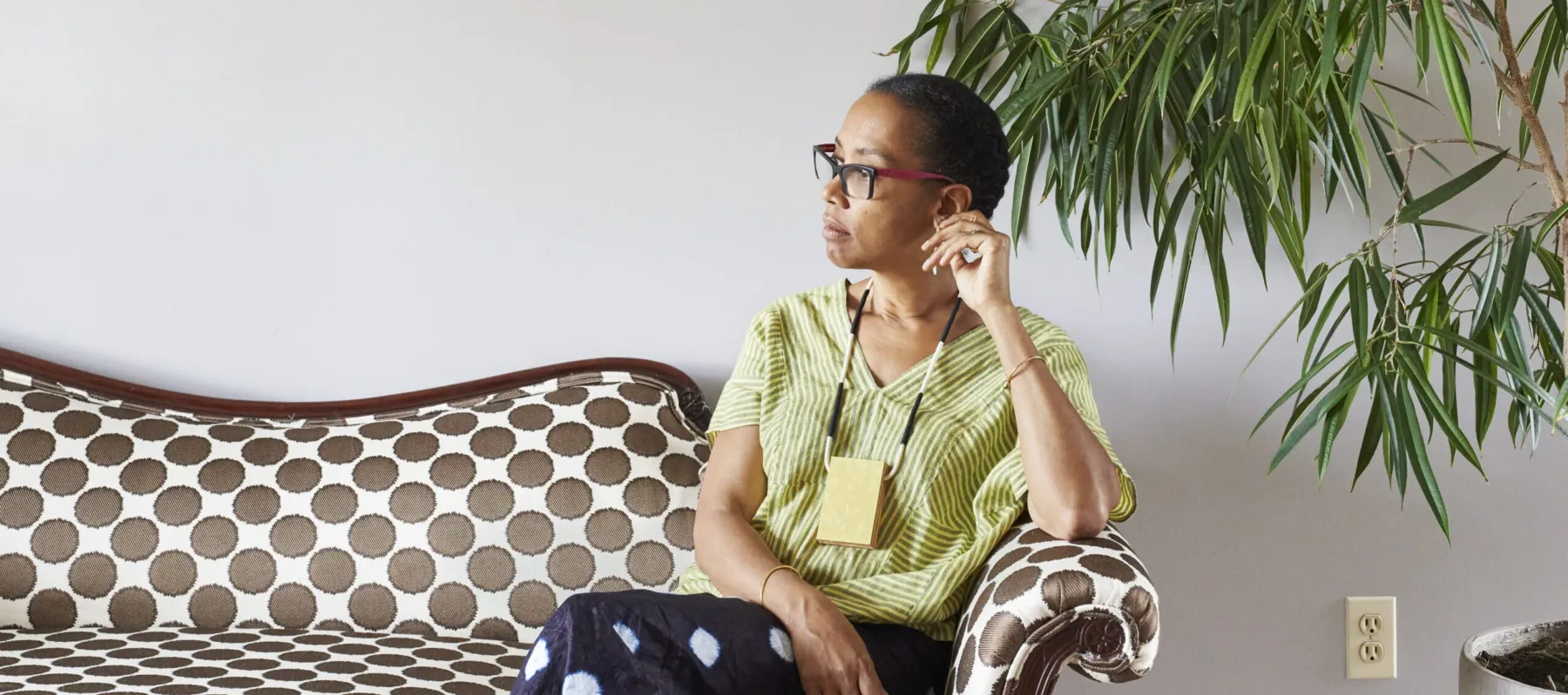 Sonya Clark calmly sits on a polka-dot couch against a white wall, next to a large plant. She is a medium-skinned adult woman with dark hair pulled back, looking to her right with her legs crossed. She wears a striped top, black pants with large white dots, glasses, and a necklace.