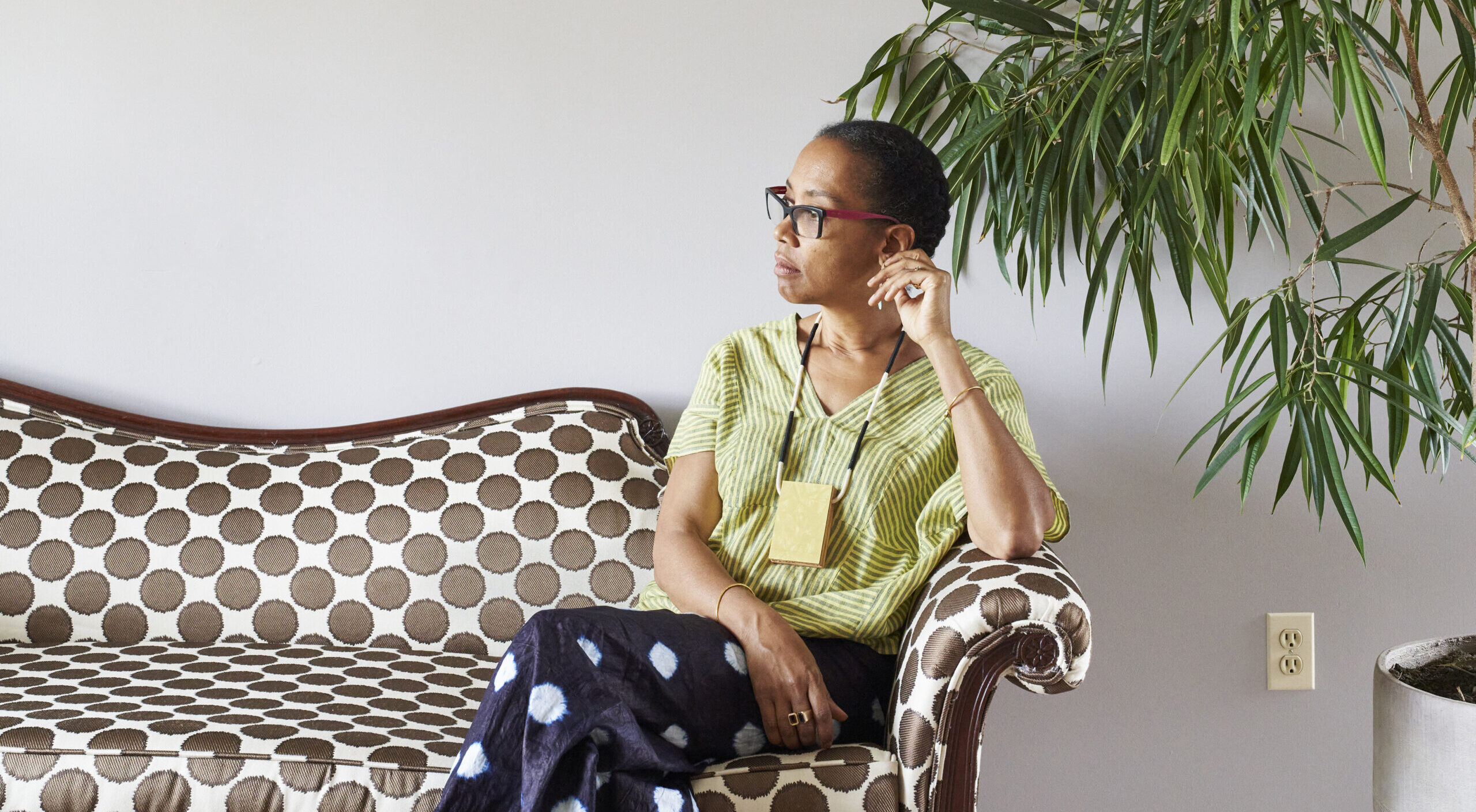 Sonya Clark calmly sits on a polka-dot couch against a white wall, next to a large plant. She is a medium-skinned adult woman with dark hair pulled back, looking to her right with her legs crossed. She wears a striped top, black pants with large white dots, glasses, and a necklace.