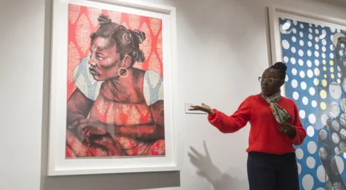 The artist, a woman with dark skin tone, speaks in front of her artwork, gesturing to it, a large colorful print of a woman with a pattern overlay across the paper. The artist wears a bright right sweater, a multicolored scarf, glasses, and gold hoops.