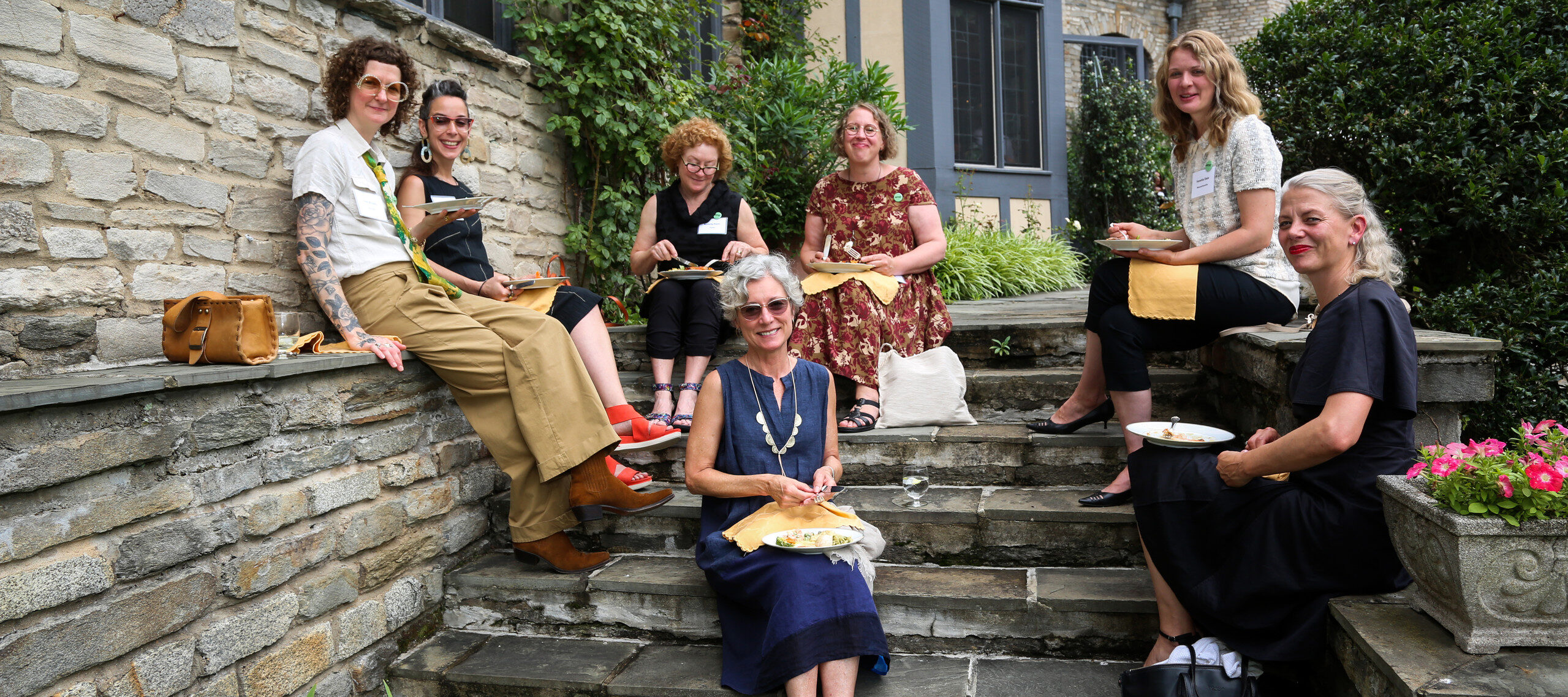 A photograph of seven light skinned women seated on stone steps eating lunch with plates and napkins in their laps. The women are looking at the camera and smiling. There is a Tudor style home in the background.