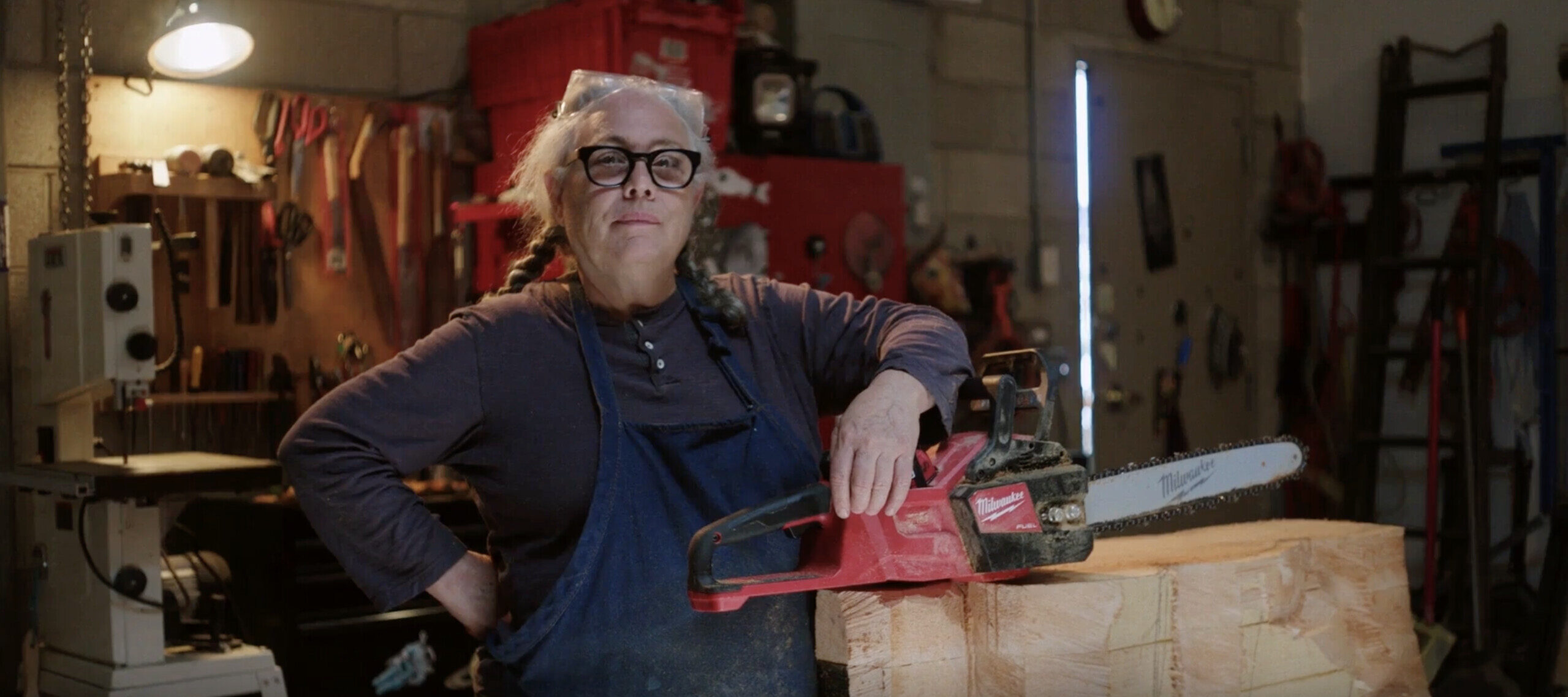A woman with medium-light skin tone, gray pigtails, large black glasses, a dark long-sleeve shirt and a navy apron stands proudly looking at the camera in a wood shop artist studio. Both her arms are bent, one at her waist and the other leaning on a chainsaw that rests on a large piece of wood on a table.