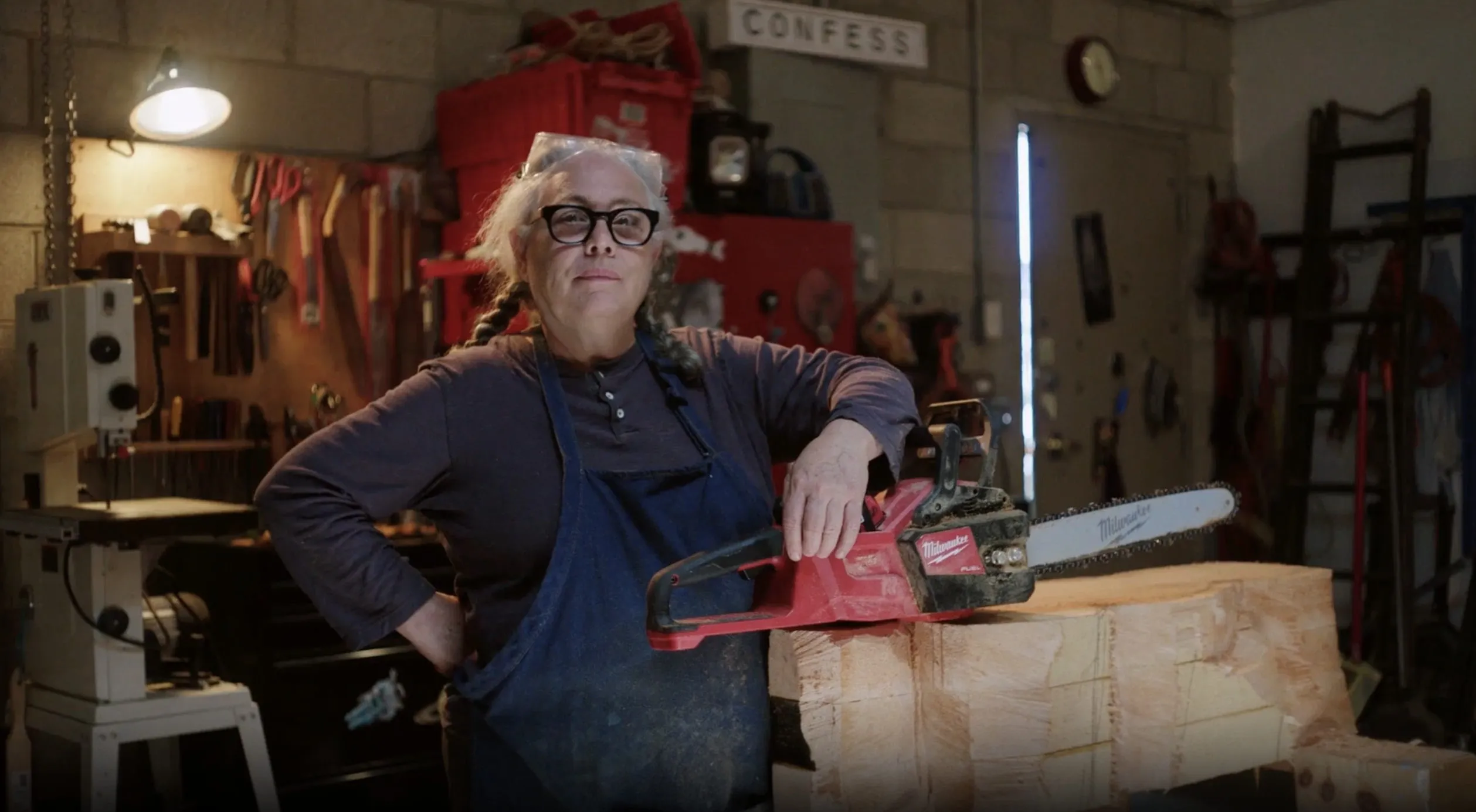 A woman with medium-light skin tone, gray pigtails, large black glasses, a dark long-sleeve shirt and a navy apron stands proudly looking at the camera in a wood shop artist studio. Both her arms are bent, one at her waist and the other leaning on a chainsaw that rests on a large piece of wood on a table.