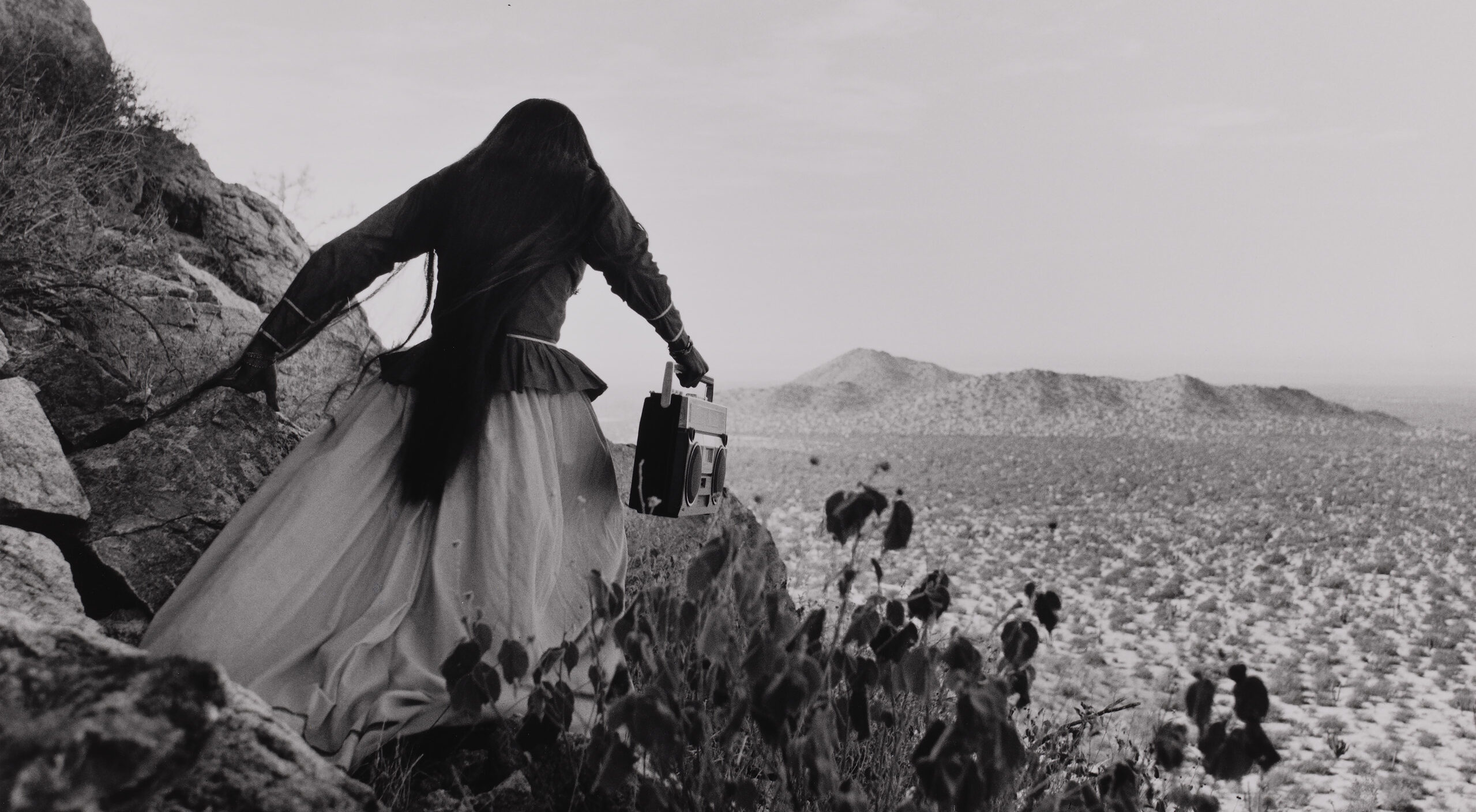 A black-and-white photograph shows the back of a woman as she crests a rocky path above a vast desert landscape beneath an expansive sky. Her traditional, ethnic full skirt, long-sleeved blouse, and long, straight, dark hair contrasts with the modern portable stereo she carries.