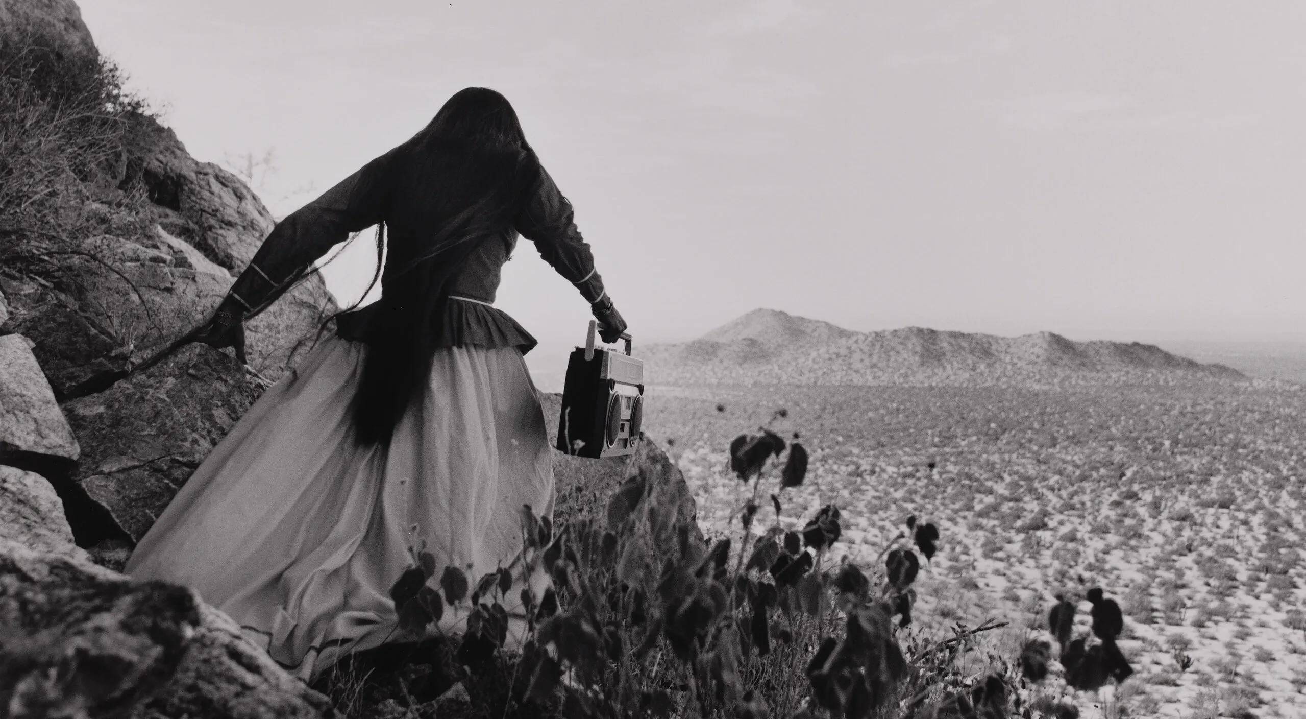 A black-and-white photograph shows the back of a woman as she crests a rocky path above a vast desert landscape beneath an expansive sky. Her traditional, ethnic full skirt, long-sleeved blouse, and long, straight, dark hair contrasts with the modern portable stereo she carries.