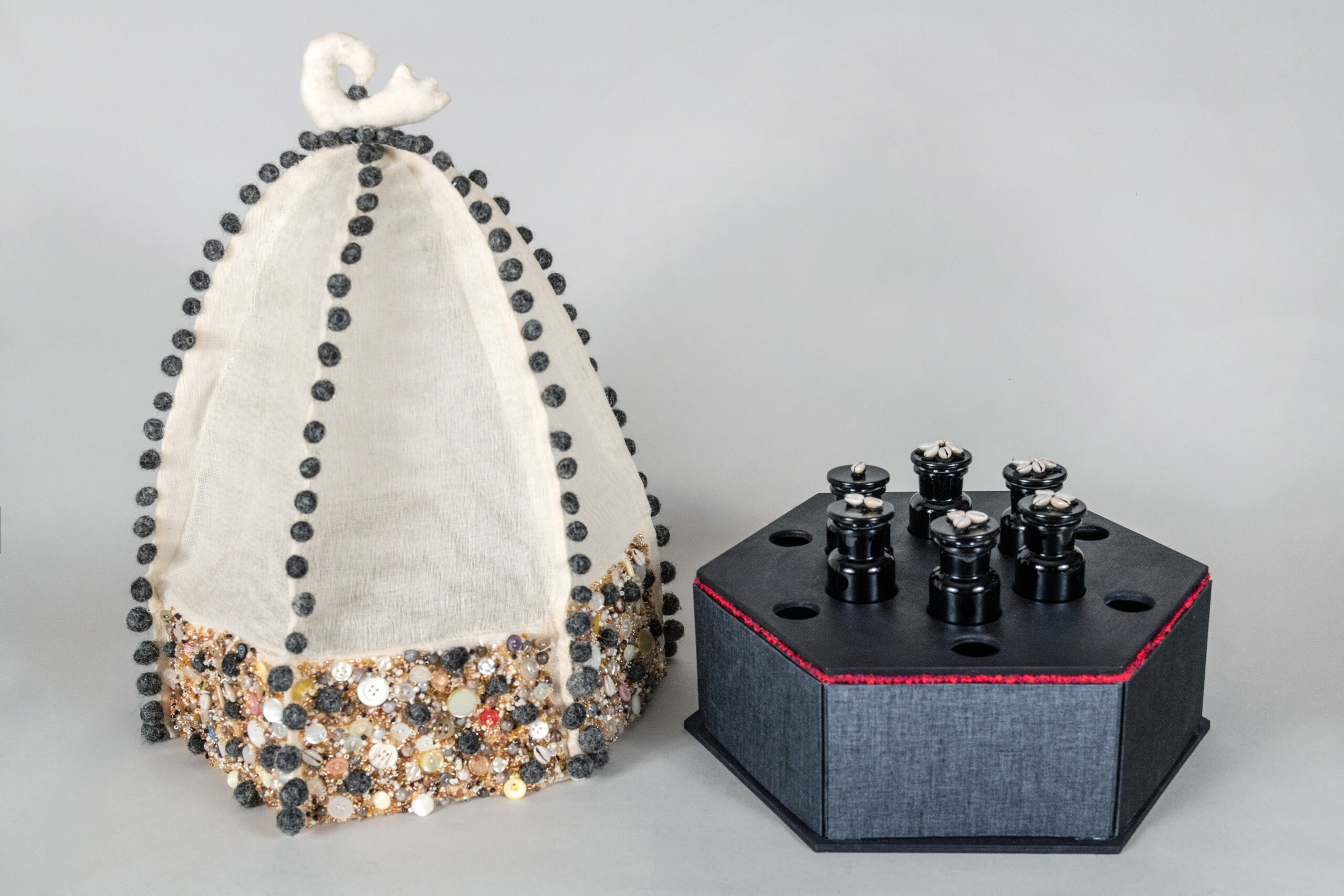 Two parts of an artist's book. One part is a black hexagonal box covered in cowry shells, small black bottles, and red beads. The other part is made of white fabric and covered in balls of human hair, buttons, and beads.