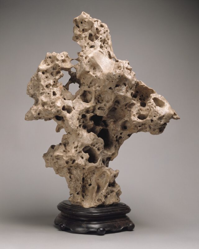 An irregular-shaped, sandy-toned stone object stands on a rounded base made of dark wood. The stone is an evocative, abstract form with rough curves, concavities, holes, and pores.