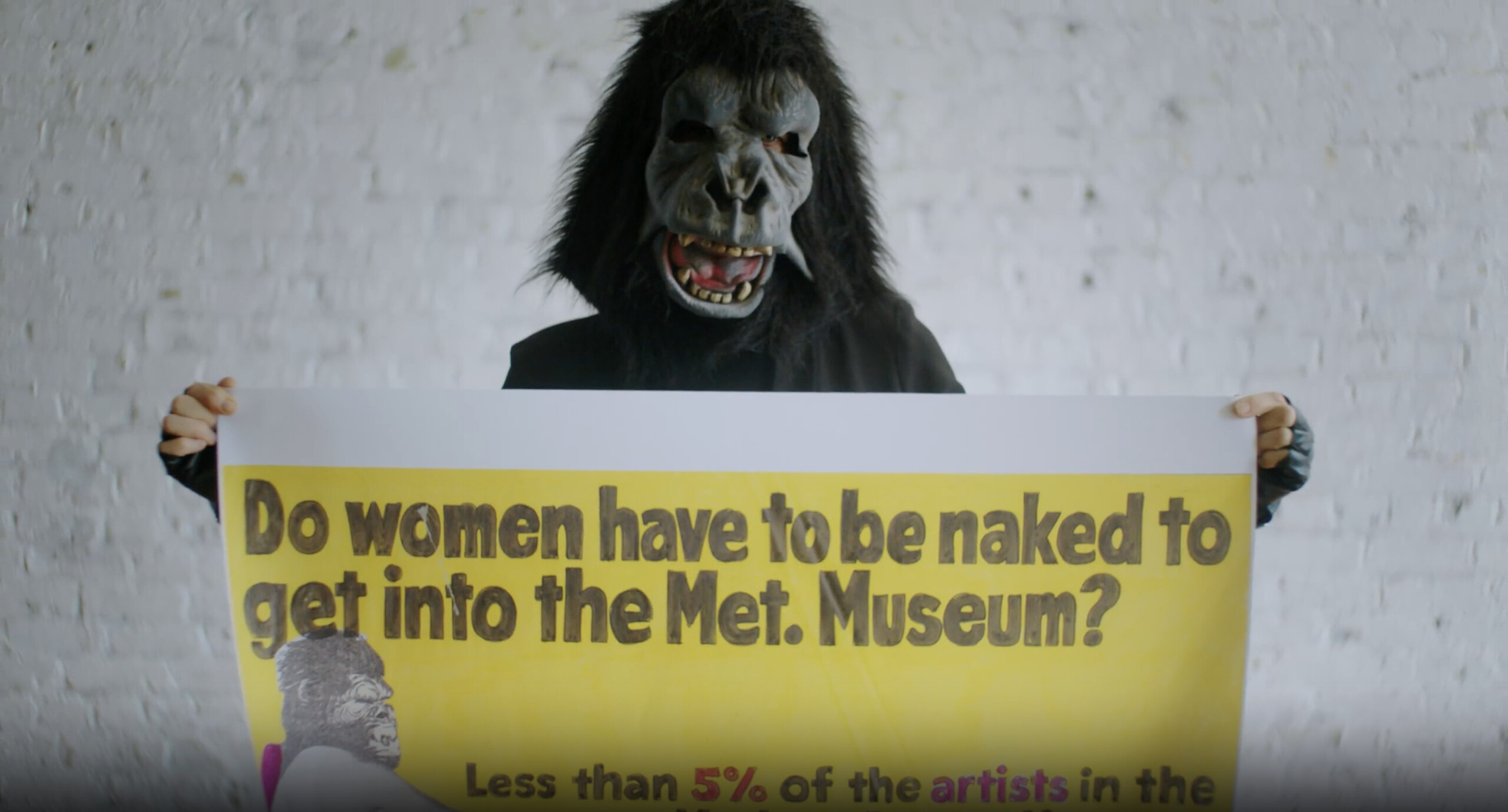 A person wearing a gorilla mask an a black long sleeve shirt holds a large poster that reads "Do women have to be naked to get into the Met. Museum?" standing against a whitewashed brick wall.