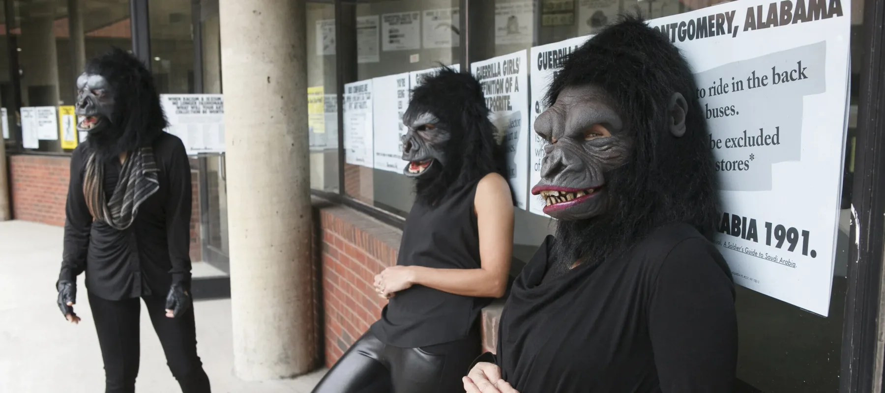 Three women dressed in all black with gorilla masks over their heads are leaning against a brick wall with a window. The window is covered in posters with big black letters that read "Guerilla Girls", among other text.