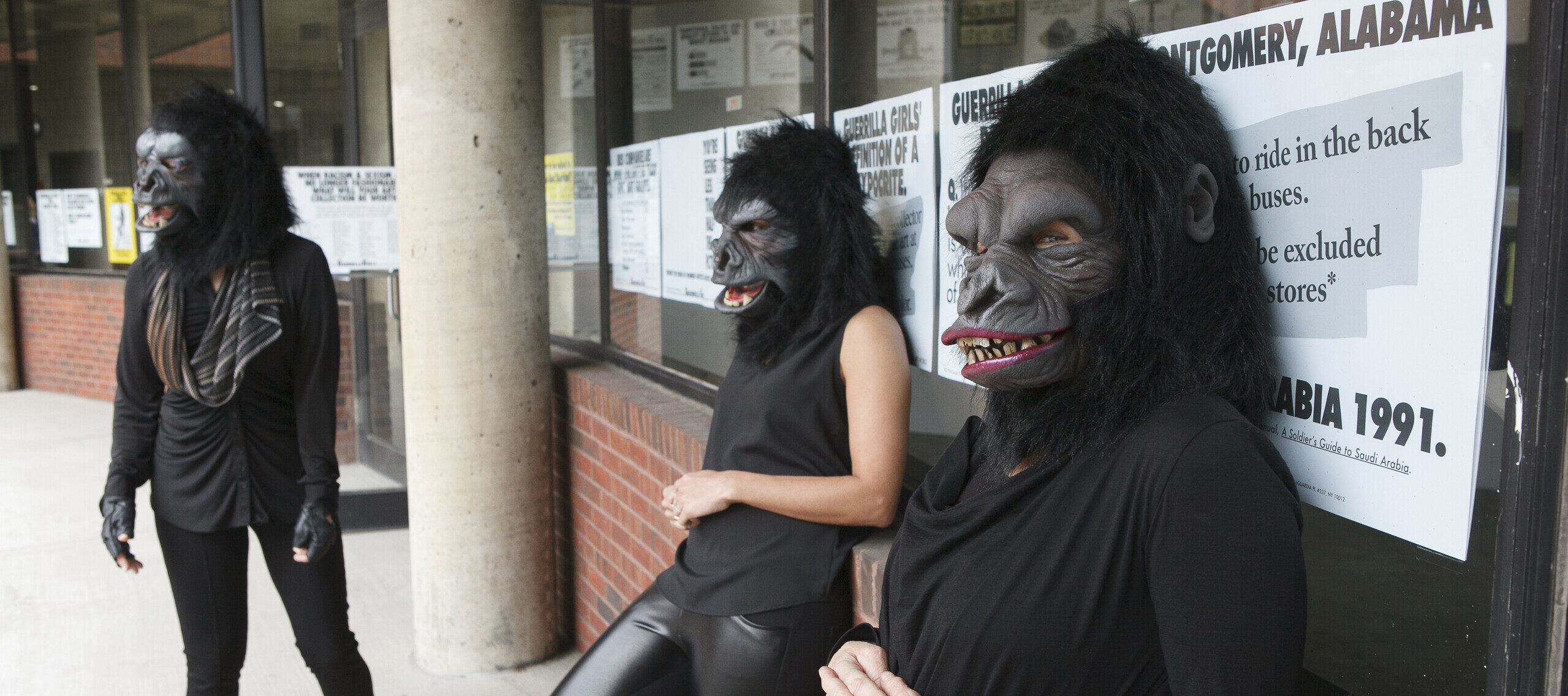 Three women dressed in all black with gorilla masks over their heads are leaning against a brick wall with a window. The window is covered in posters with big black letters that read 