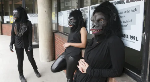 Three women dressed in all black with gorilla masks over their heads are leaning against a brick wall with a window. The window is covered in posters with big black letters that read "Guerilla Girls", among other text.