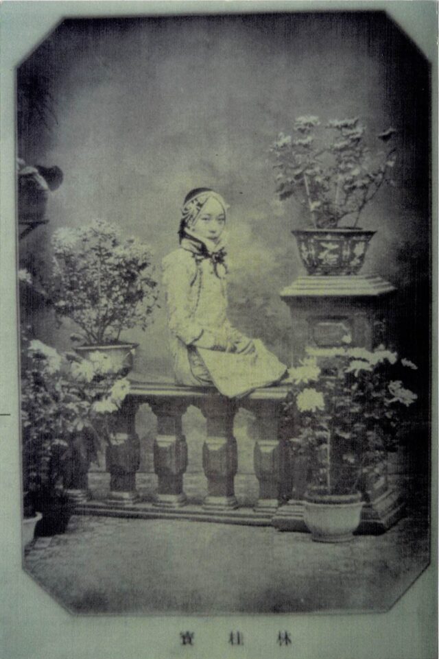 In this black-and-white photo, a young woman of Asian descent sits on a balustrade surrounded by potted plants. Her hands are folded on her lap, and she wears light clothing and a headpiece.