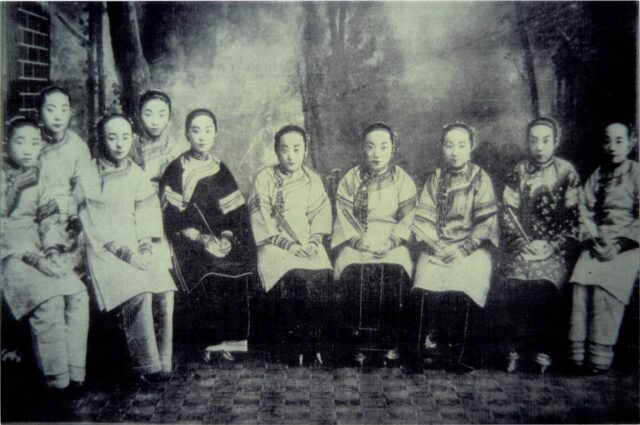 A black-and-white image shows a group of Asian women in traditional Chinese attire. They are seated facing the camera, with their hands folded on their laps.