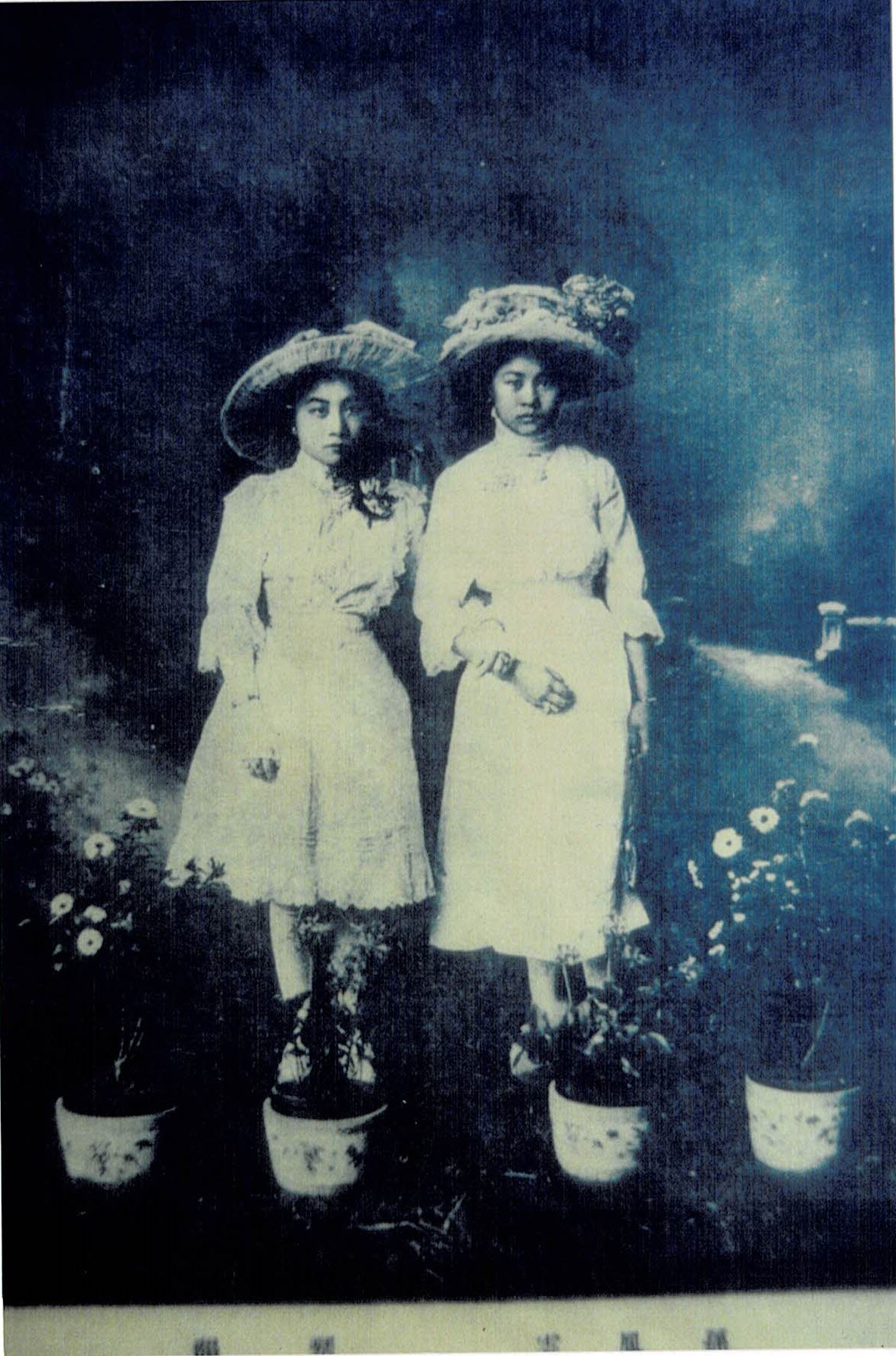 A black-and-white image shows two young girls wearing white Western dresses and large flowered hats. They stand with linked arms in a setting with four small flowerpots on the ground.
