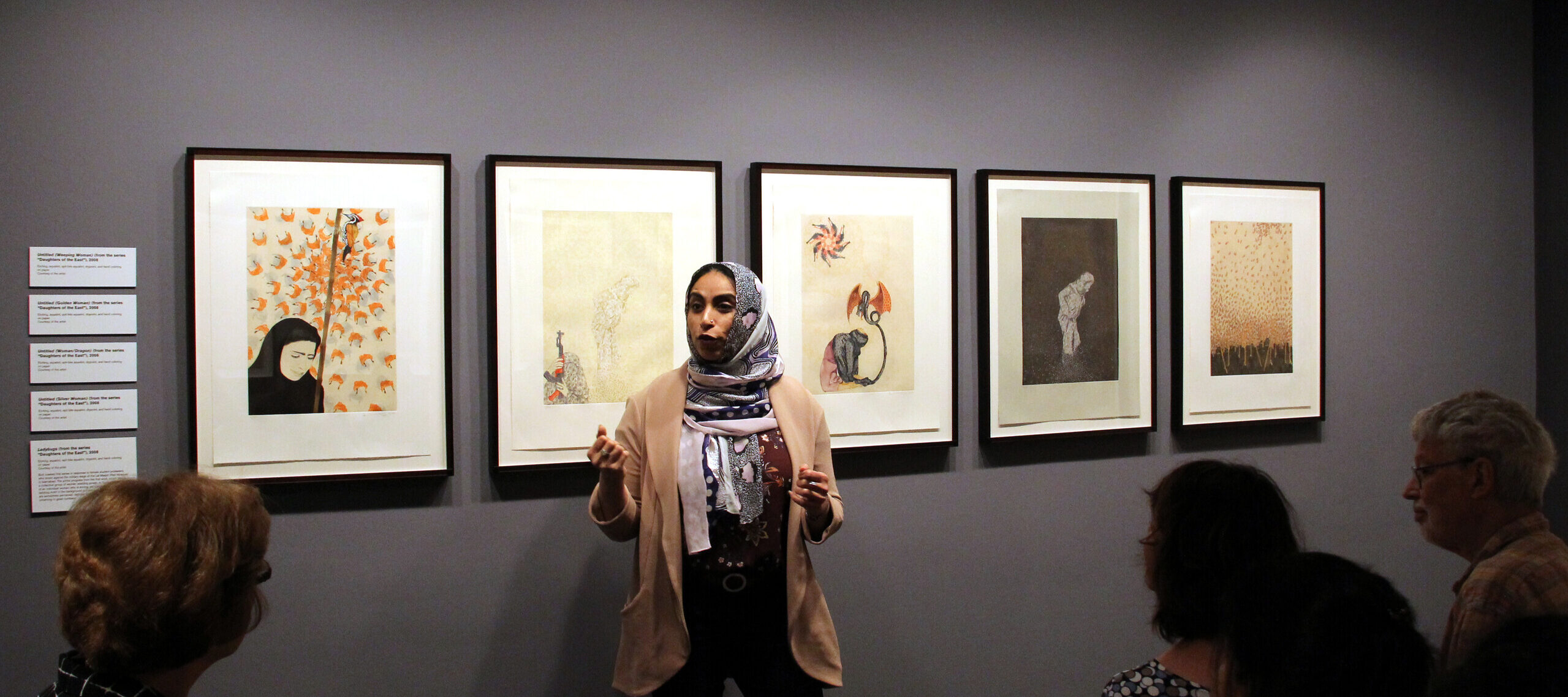 A woman with medium skin tone wearing a multicolored head scarf, beige cardigan, maroon top, and black pants stands in a gallery speaking in front of group of people sitting and listening. The speaker is the artist gesturing while speaking in front of five intricate, detailed prints.