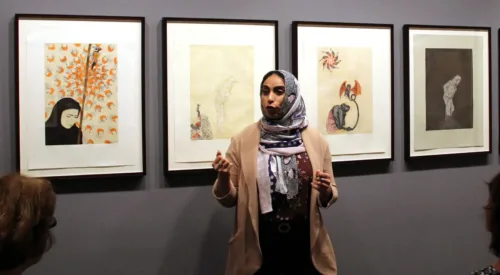 A woman with medium skin tone wearing a multicolored head scarf, beige cardigan, maroon top, and black pants stands in a gallery speaking in front of group of people sitting and listening. The speaker is the artist gesturing while speaking in front of five intricate, detailed prints.