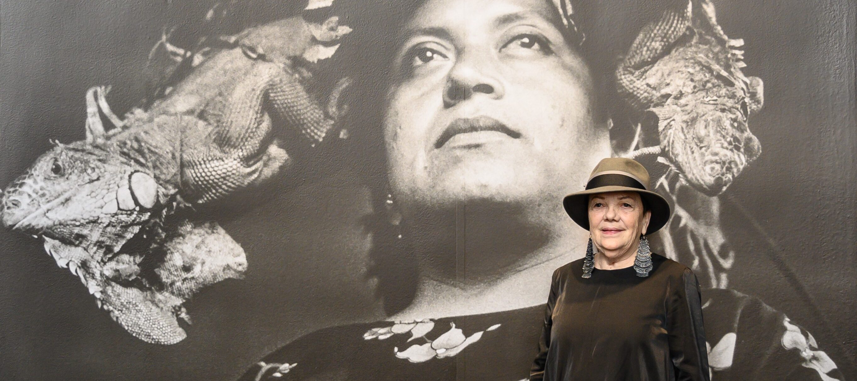 A woman in a fedora and a long black dress poses in front of a large black-and-white photograph of a woman wearing a floral dress and a large headdress who is standing, looking upward.