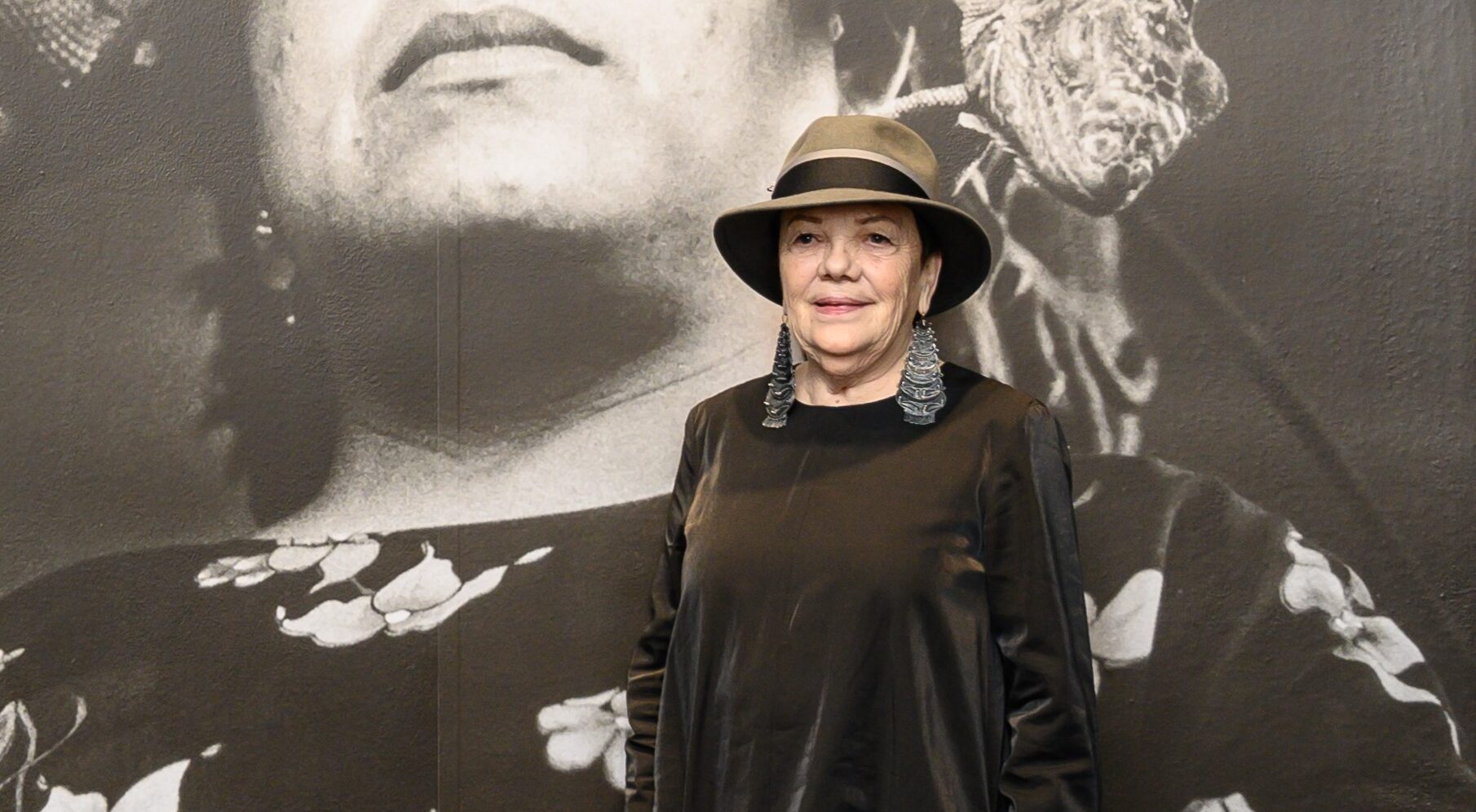 A woman in a fedora and a long black dress poses in front of a large black-and-white photograph of a woman wearing a floral dress and a large headdress who is standing, looking upward.