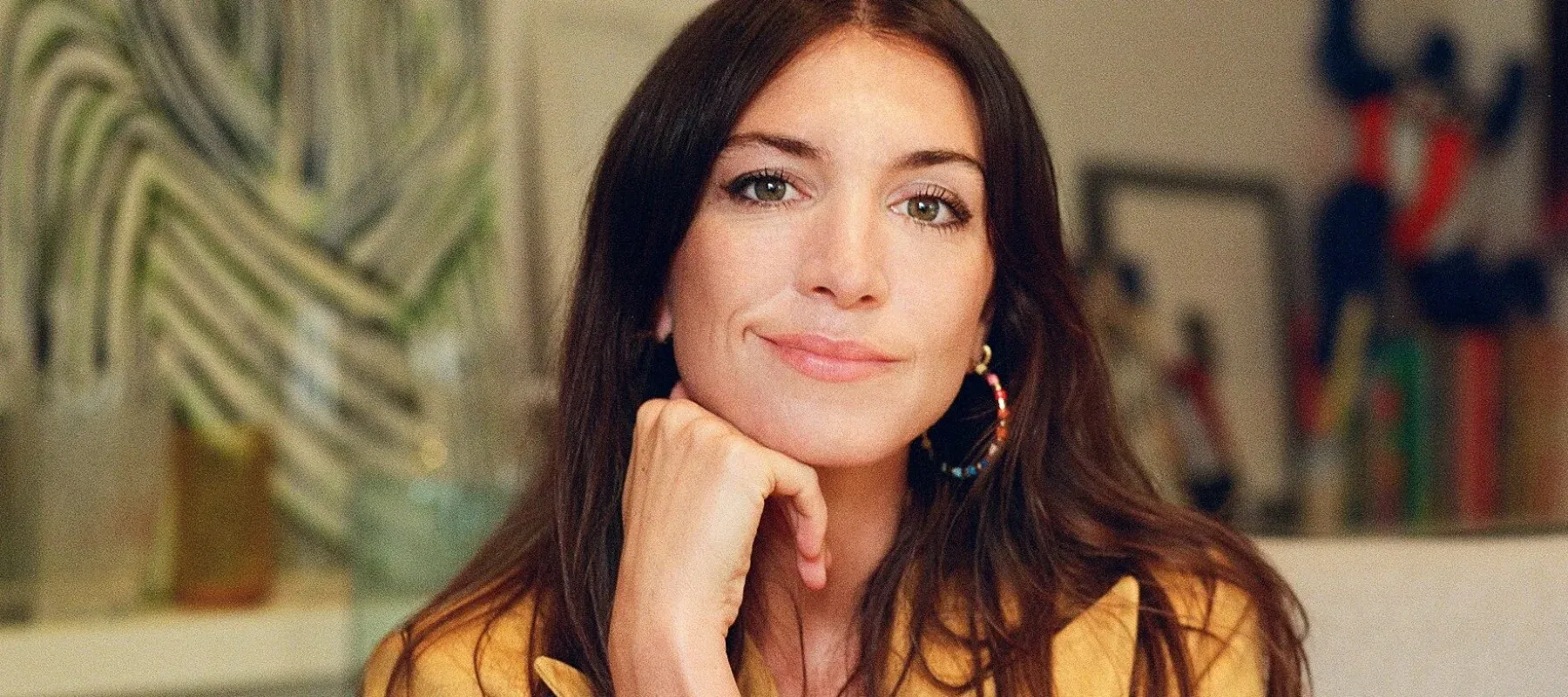 Photograph of an adult woman, in a yellow suit, sitting in a chair with artwork behind her.