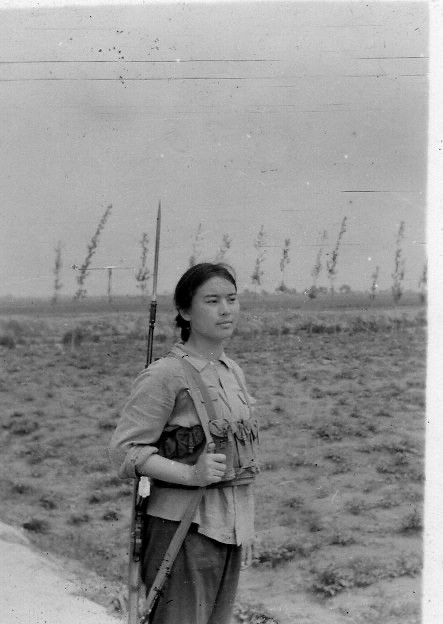 A black-and-white image shows a young woman with dark hair in a braid standing in an empty field. She wears a button-up shirt, trousers, and a vest of tactical gear. She gazes into the distance and has a rifle slung over her shoulder.