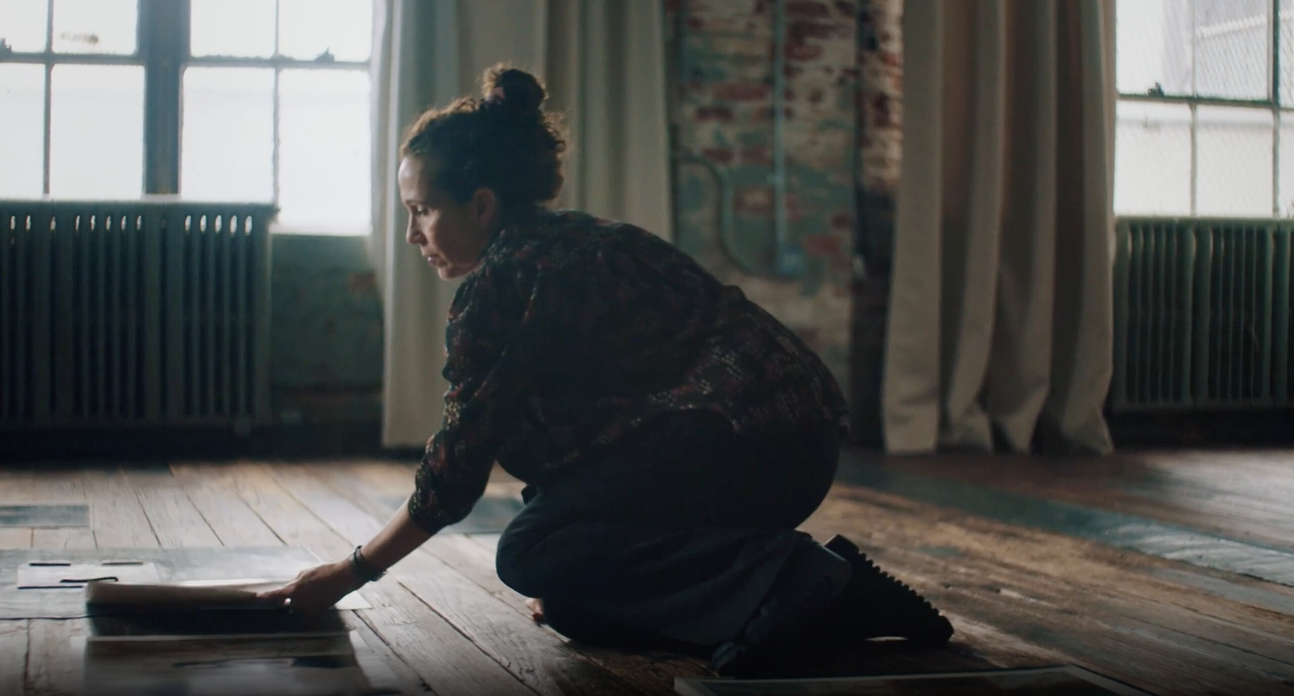 A woman with medium skin tone and a loose top-knot bun kneels on the floor leaning forward as she lays a photograph on the wooden floor. She is in a large room with big windows and exposed brick walls.