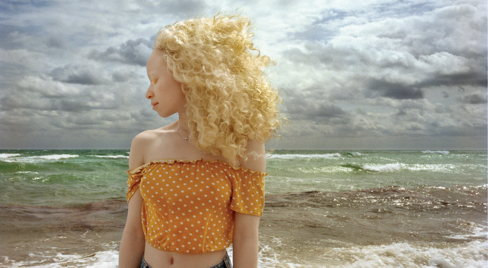 In front of a stormy ocean, a woman with light skin and blonde, wind-blown hair stands with head turned in profile and eyes closed.