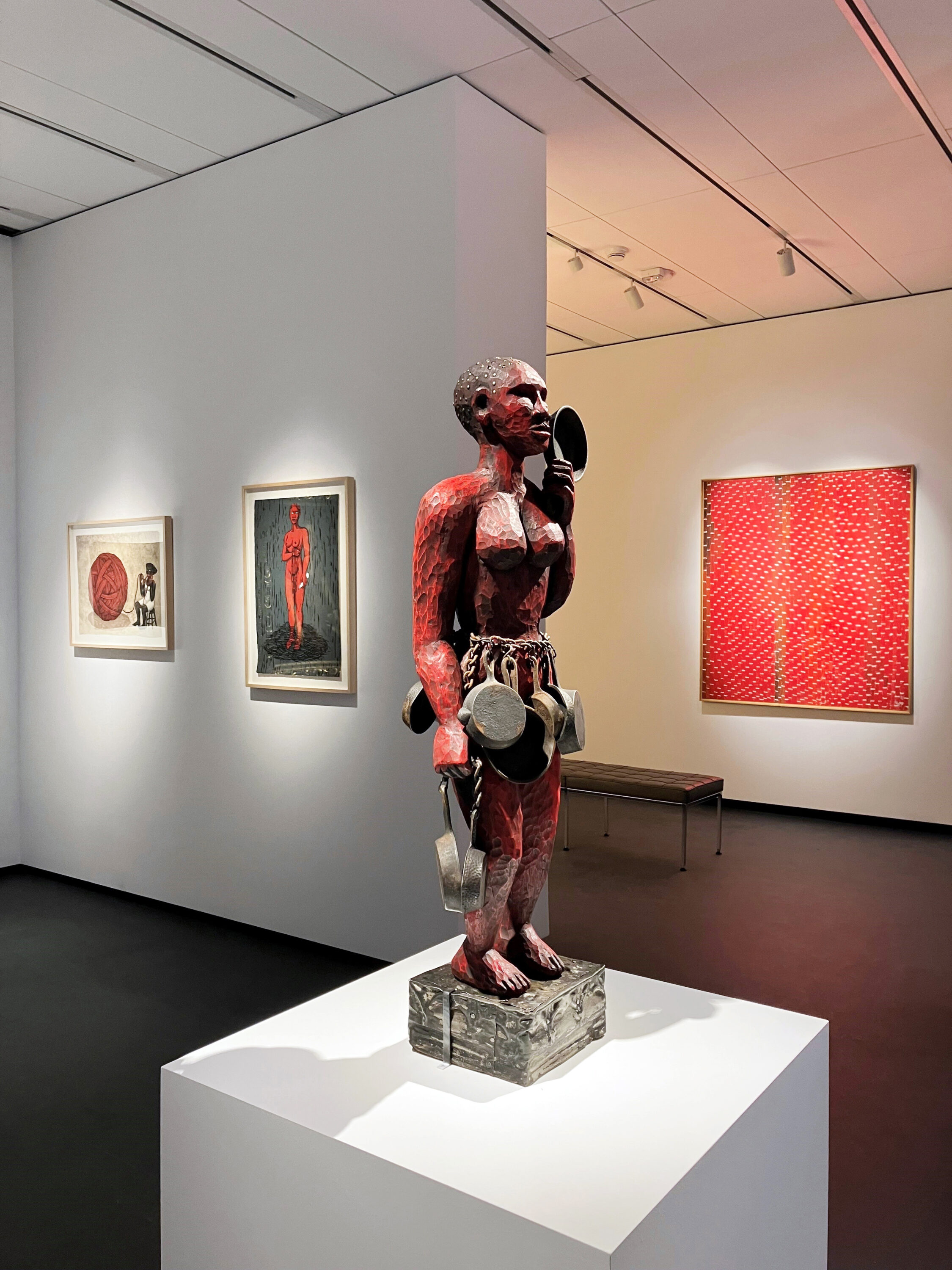 Artworks featuring the color red are displayed in a modern museum gallery. A central carved wooden sculpture of a powerful figure painted red and holding cast iron skillets is flanked by a vibrant red abstract painting and two prints that highlight the color.