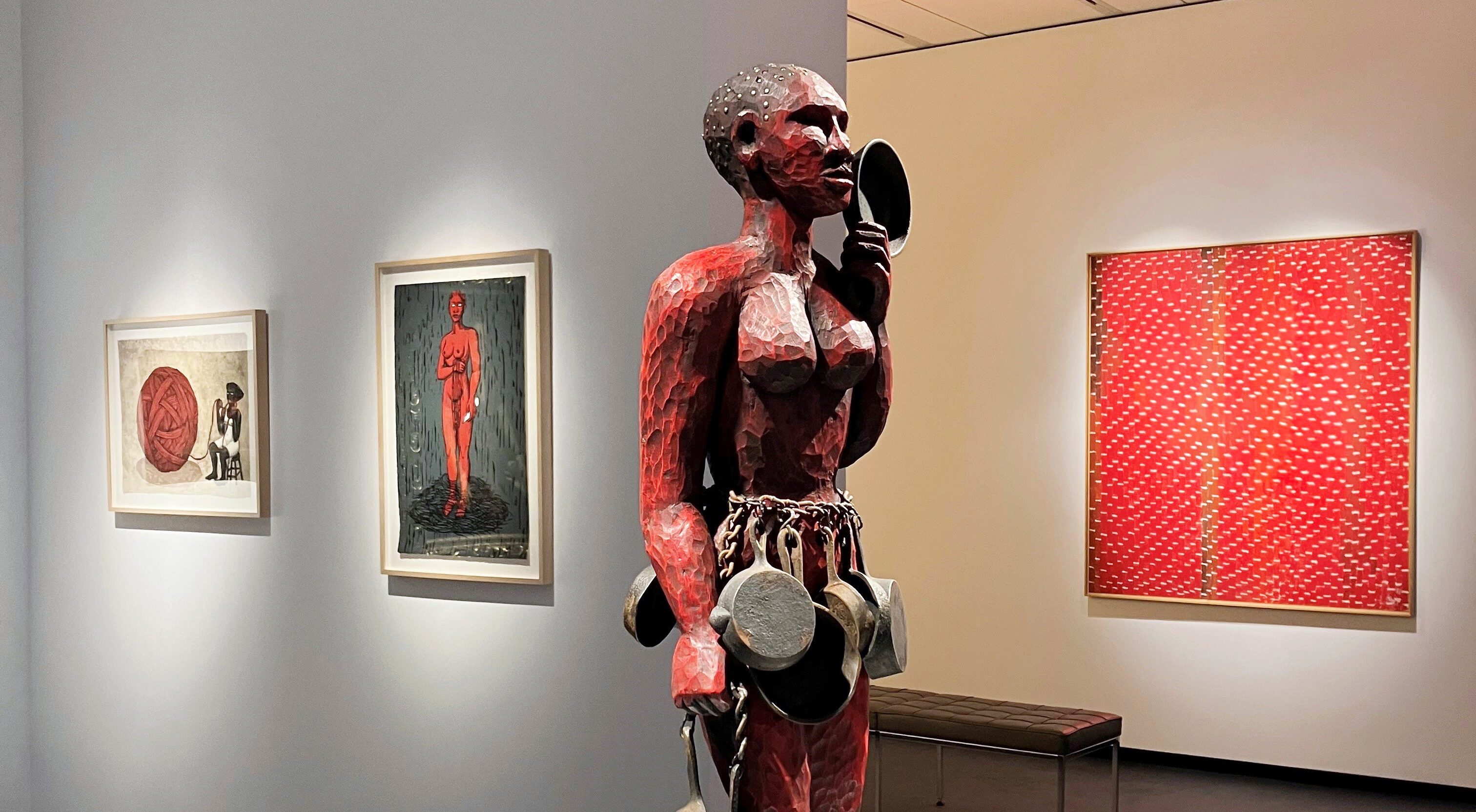 Artworks featuring the color red are displayed in a modern museum gallery. A central carved wooden sculpture of a powerful figure painted red and holding cast iron skillets is flanked by a vibrant red abstract painting and two prints that highlight the color.