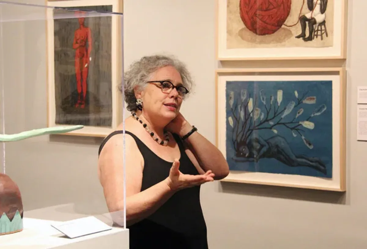 A light-skinned, adult woman speaking with one hand extended and the other touching her neck in a white gallery filled with her colorful artworks depicting African-American women. She has curly gray hair, glasses, and wears a simple, black, sleeveless top and a necklace.