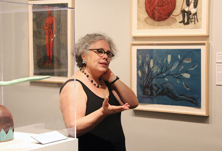 A light-skinned, adult woman speaking with one hand extended and the other touching her neck in a white gallery filled with her colorful artworks depicting African-American women. She has curly gray hair, glasses, and wears a simple, black, sleeveless top and a necklace.