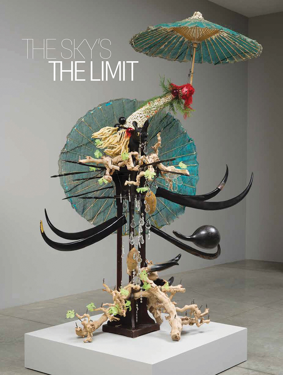 A book cover features the image of a sculpture on a white base. It is assembled from many materials: blue parasols, black wooden horns, a black wooden rhino figurine, green nylon, beaded flowers, a red doll’s head, clear glass chandelier drops, and more. Just above it, white text reads “The Sky’s the Limit” in all caps.