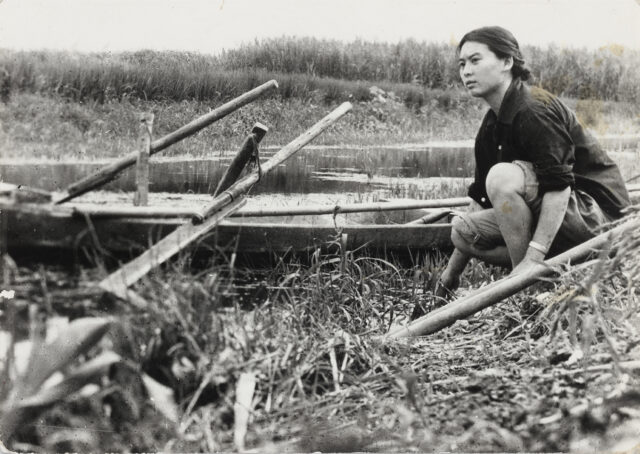 A black-and-white image shows a young Asian woman crouching in a rural setting, on the bank of a river where a rowboat rests on the water. She stares into the distance.