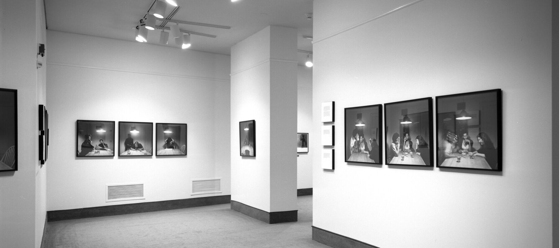 A view of a gallery space. A sequence of photographs of a woman with a dark skin tone sitting around a kitchen table is hanging on the walls. On each photograph, different people join her at the table.