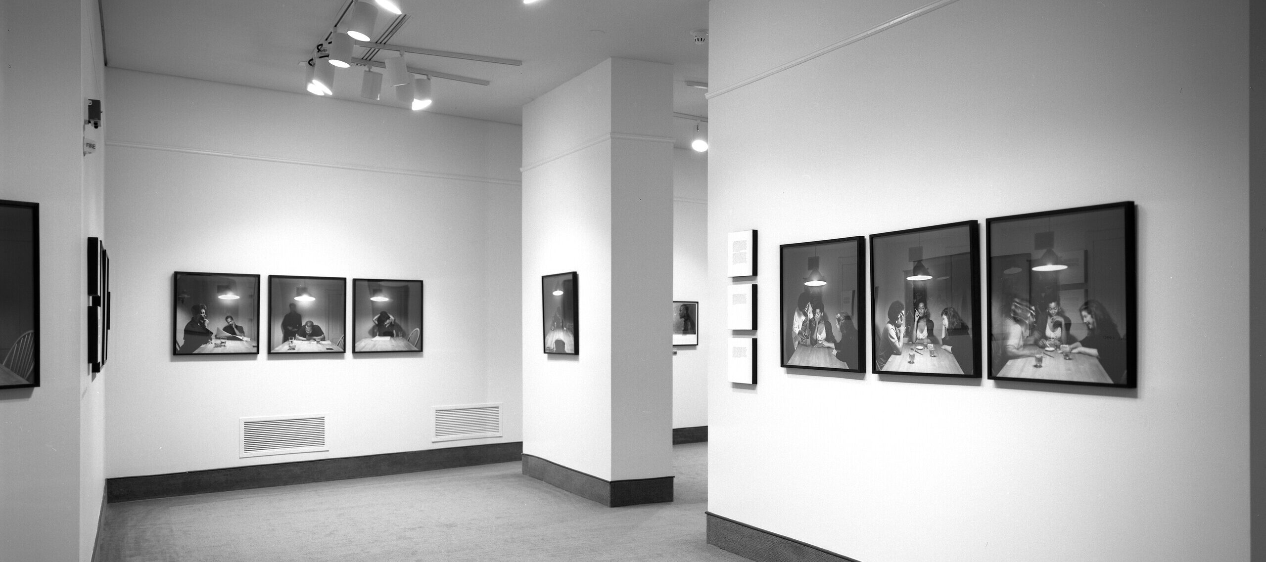 A view of a gallery space. A sequence of photographs of a woman with a dark skin tone sitting around a kitchen table is hanging on the walls. On each photograph, different people join her at the table.