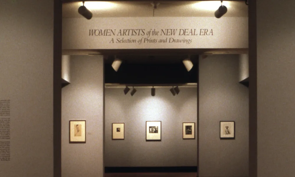 A view of a gallery space. The walls are painted gray, and several artworks are hanging on them. In large letters, it says on a wall "Women Artists of the New Deal Era: A Selection of Prints and Drawings."