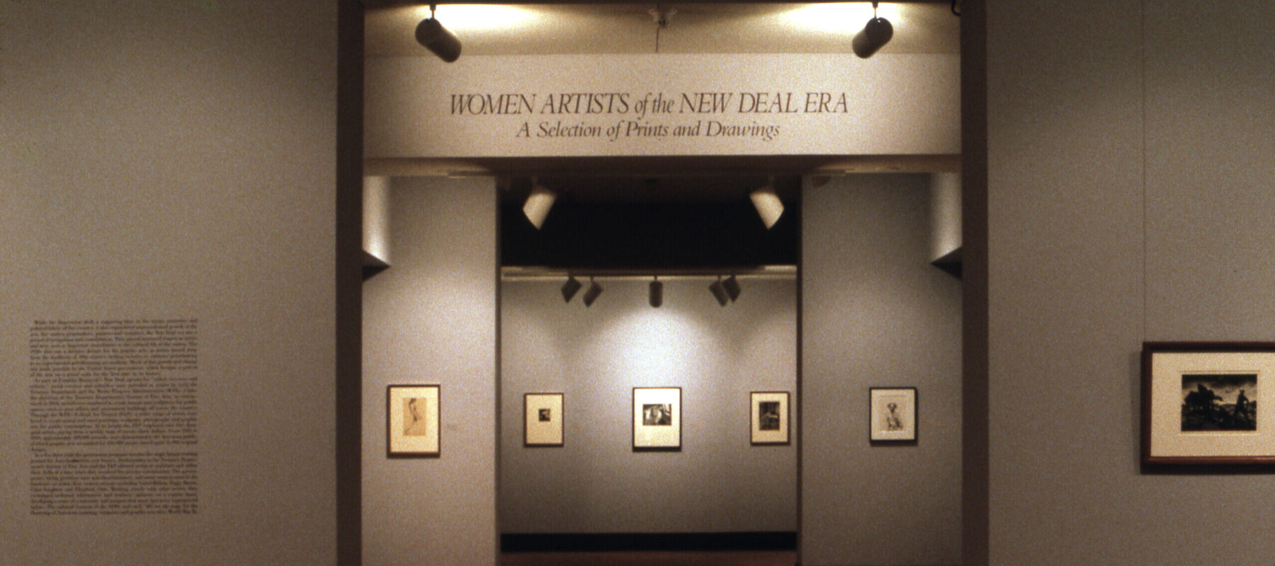 A view of a gallery space. The walls are painted gray, and several artworks are hanging on them. In large letters, it says on a wall "Women Artists of the New Deal Era: A Selection of Prints and Drawings."