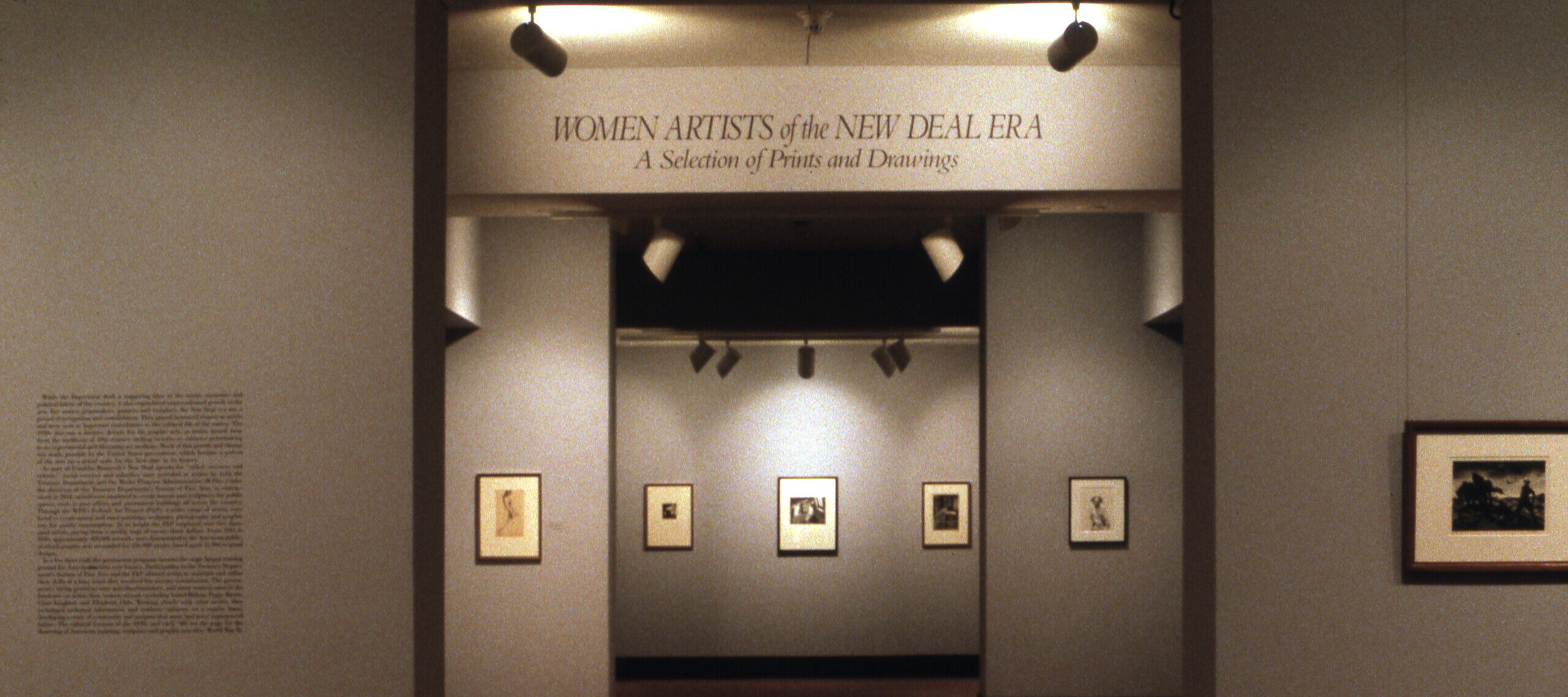 A view of a gallery space. The walls are painted gray, and several artworks are hanging on them. In large letters, it says on a wall 