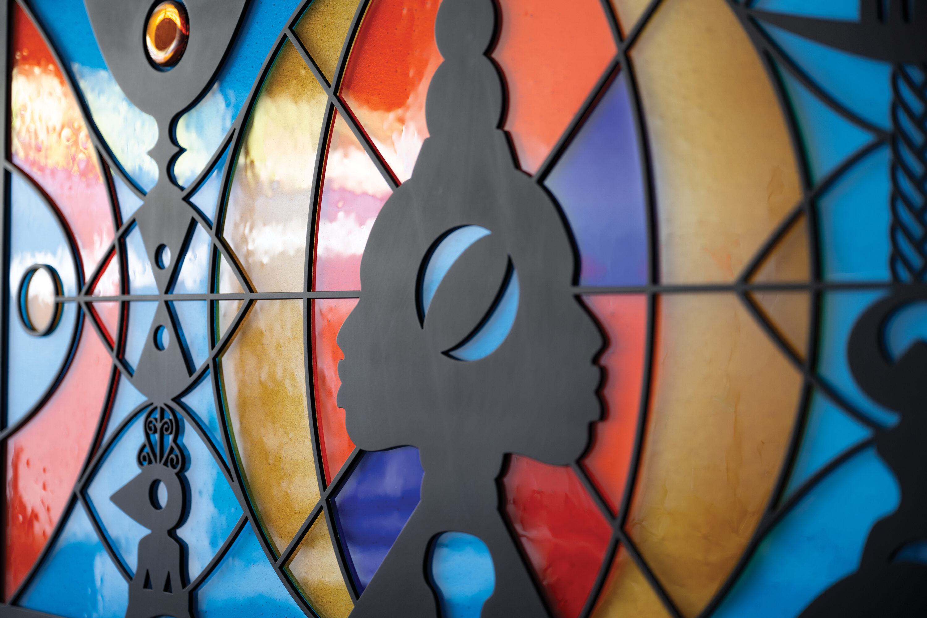A sculpture hangs flat against the wall. It looks like a wide, stained glass window that can be lit from behind. There are two faces in profile in the center, positioned back-to-back, as if emerging from a line down the center of the piece. They are made out of black metal, as are several totem-like designs positioned in front of bright orange, blue-green, and yellow abstract shapes. The abstract shapes are made of fused glass.
