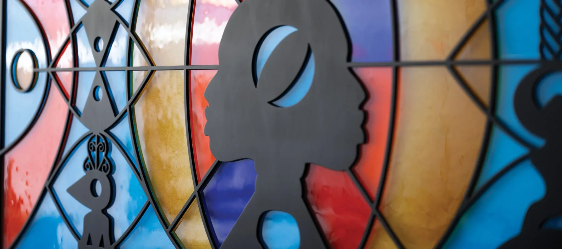 A sculpture hangs flat against the wall. It looks like a wide, stained glass window that can be lit from behind. There are two faces in profile in the center, positioned back-to-back, as if emerging from a line down the center of the piece. They are made out of black metal, as are several totem-like designs positioned in front of bright orange, blue-green, and yellow abstract shapes. The abstract shapes are made of fused glass.