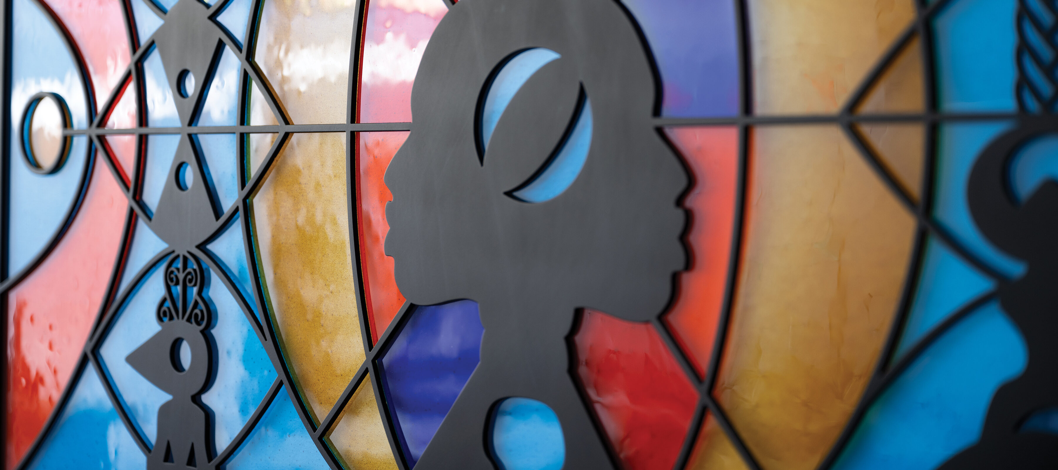 A sculpture hangs flat against the wall. It looks like a wide, stained glass window that can be lit from behind. There are two faces in profile in the center, positioned back-to-back, as if emerging from a line down the center of the piece. They are made out of black metal, as are several totem-like designs positioned in front of bright orange, blue-green, and yellow abstract shapes. The abstract shapes are made of fused glass.