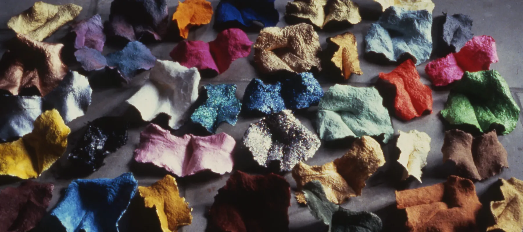 A cluster of homemade paper fragments in a variety of saturated colors sit on a tile floor. Some fragments are matte and some have glitter embedded in the paper.