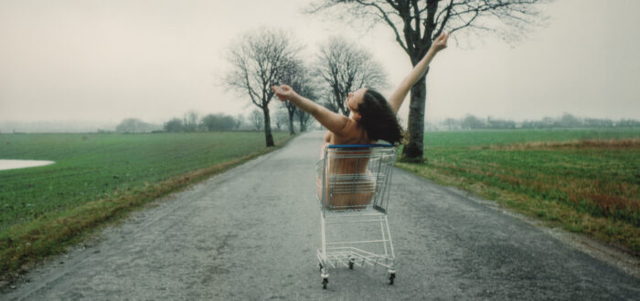 A photograph shows the nude artist sitting in a metal grocery cart. It is located on a paved road in a flat, empty landscape under a gray, misty sky. Her back to the viewer, the light-skinned, brunette woman holds her raised arms in a wide V-shape, suggesting joy or abandon.