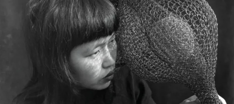 A black-and-white photograph of Ruth Asawa holding one of her large, wire crochet sculptures, draped over her shoulder and in both hands. She is a light-skinned, Asian, adult woman with black hair and blunt bangs.