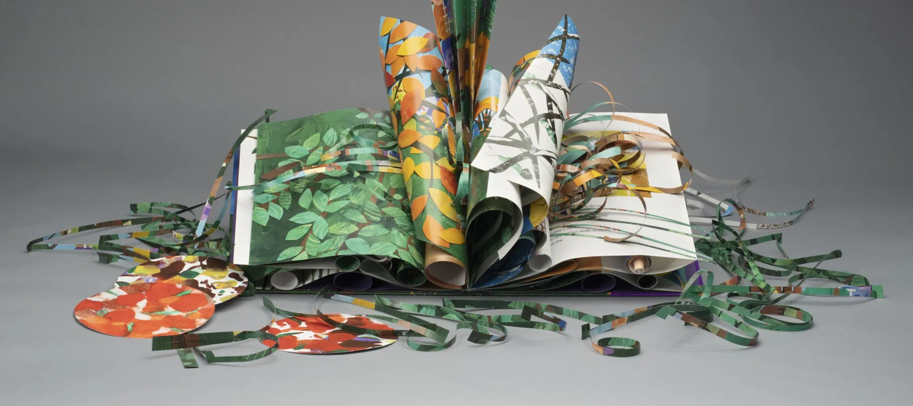 An artist's book made of colorful green, yellow, brown, orange, and red paper. Several of the pages are curled and folded. Other paper is cut into strips and circles and displayed around the book.