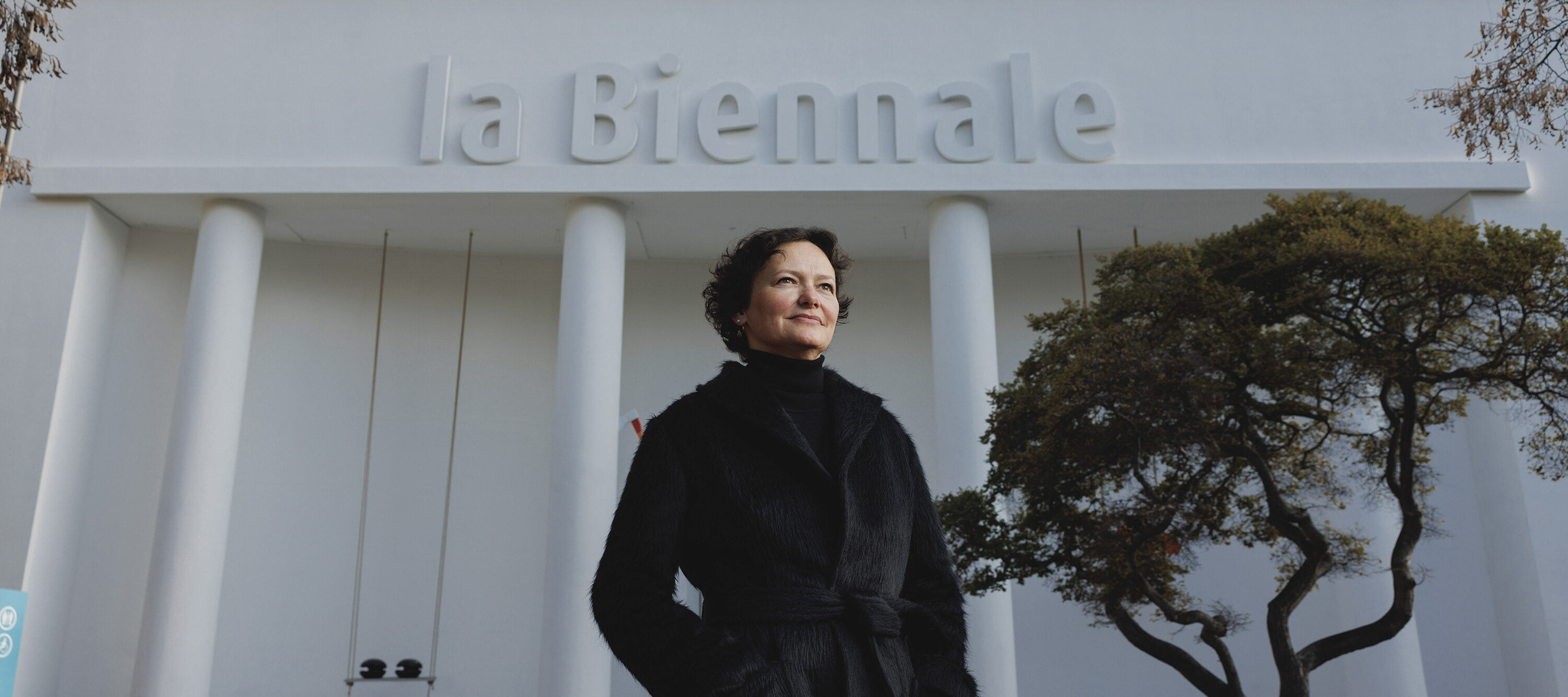 A woman in a black coat stands outside near a white building that has the name “la Biennale” on the top. She has a light skin tone and her dark brown hair is short and curly. A tree is in the background and she stares out, looking into the distance.