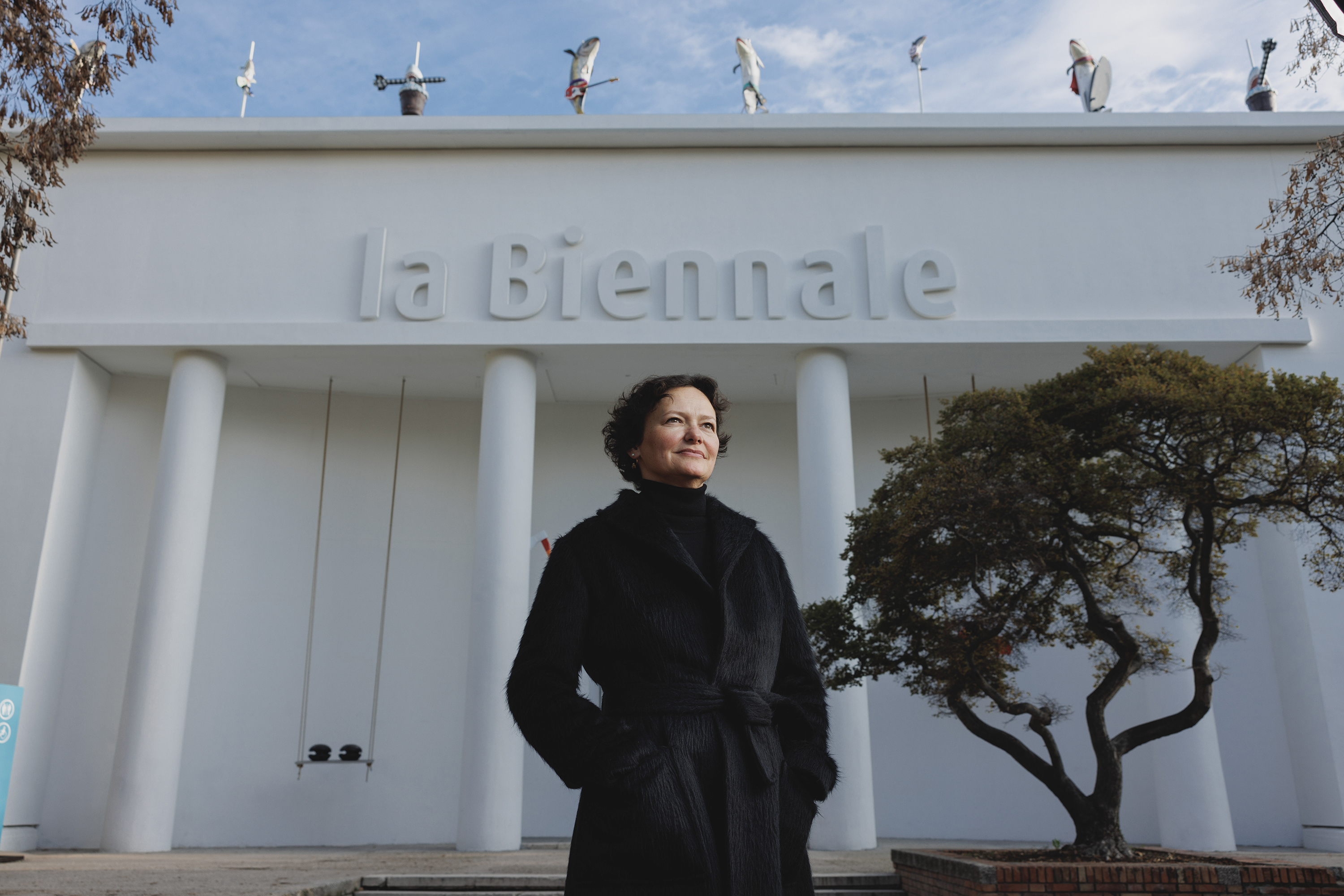 A woman in a black coat stands outside near a white building that has the name “la Biennale” on the top. She has a light skin tone and her dark brown hair is short and curly. A tree is in the background and she stares out, looking into the distance.