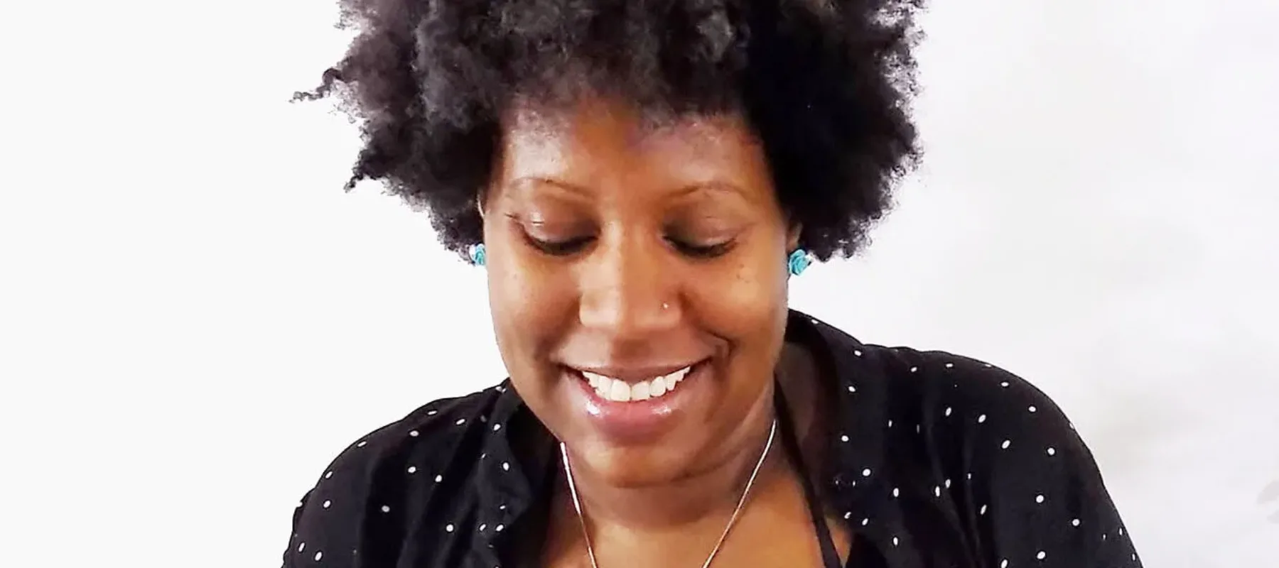 A medium-dark skinned woman with short, curly hair wears turquoise flower earrings, a nose ring, a black shirt with white dots, and an apron with a flower patch. She looks down at a small textured book she’s holding and flipping through. In the foreground are two colorful handmade books standing upright on a table.