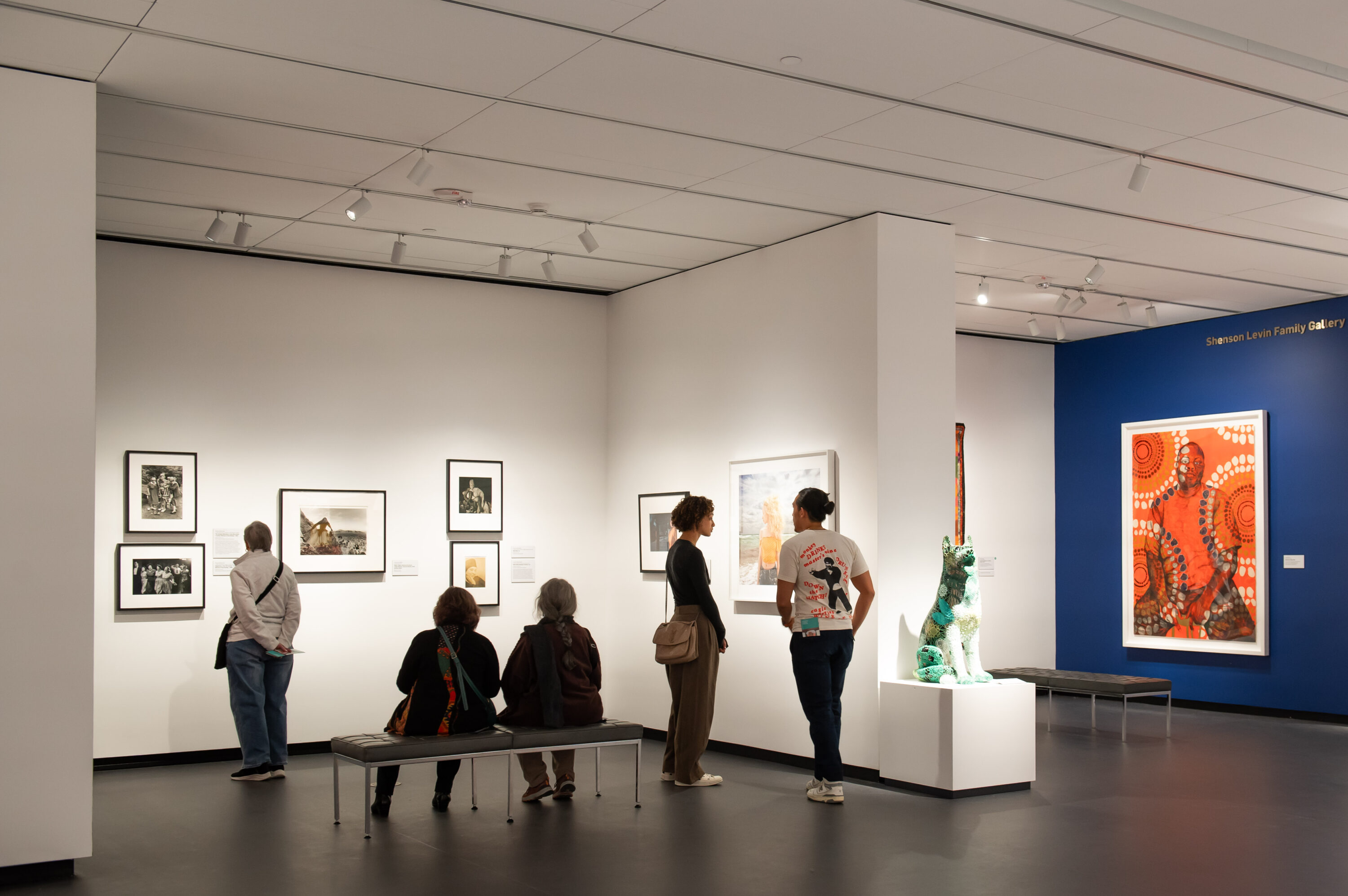 A modern museum gallery is photographed at a wide angle. Visitors observe large artworks on the walls and sculptures on pedestals.