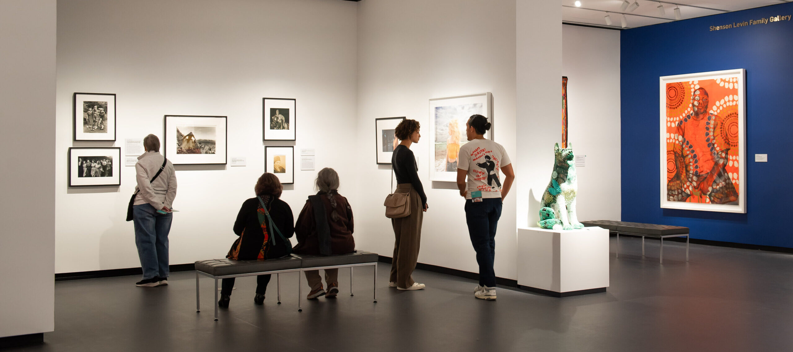 A modern museum gallery is photographed at a wide angle. Visitors observe large artworks on the walls and sculptures on pedestals.