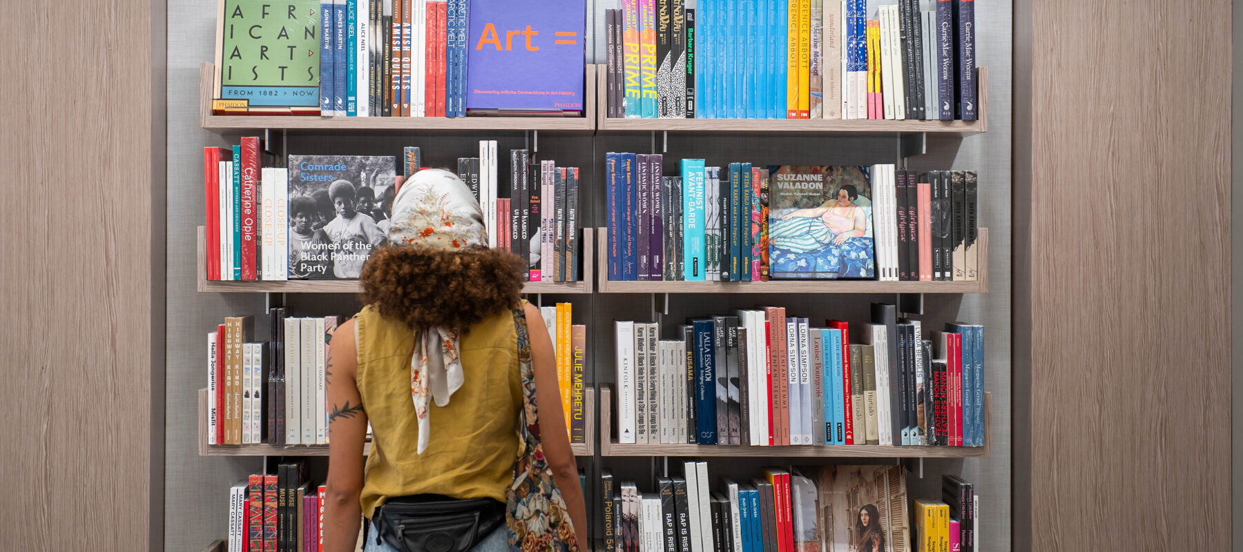 A visitor with medium skin tone and brown curly hair wearing a white and red floral head scarf, ochre sleeveless shirt, black backwards fanny pack, multicolored tote bag, and jeans stands facing a large floating bookcase filled with art books and catalogues.