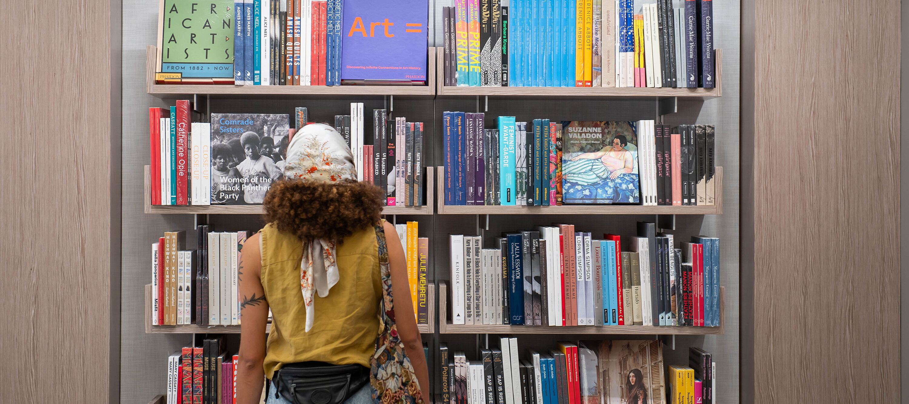A visitor with medium skin tone and brown curly hair wearing a white and red floral head scarf, ochre sleeveless shirt, black backwards fanny pack, multicolored tote bag, and jeans stands facing a large floating bookcase filled with art books and catalogues.